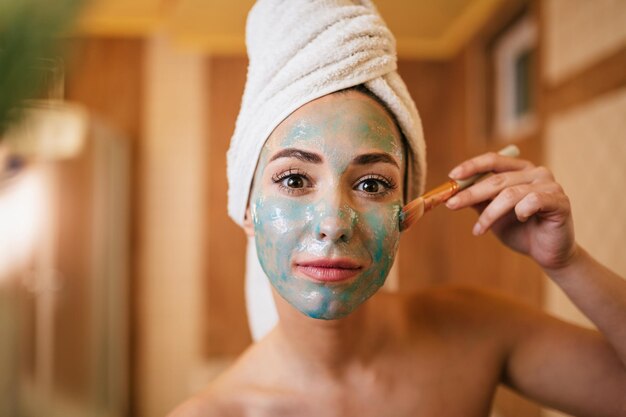 Mujer hermosa joven usando cepillo mientras aplica máscara facial en el baño