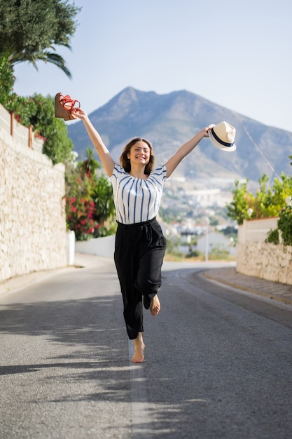 Mujer hermosa joven en vacaciones saltando. En una mano sandalias en el sombrero de segunda mano.