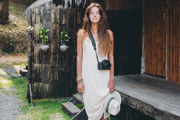 Mujer hermosa joven en vacaciones tropicales en Asia, estilo veraniego, vestido blanco boho, zapatillas, cámara de fotos digital, viajero, sombrero de paja, relajado