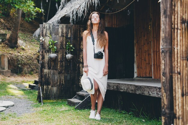 Mujer hermosa joven en vacaciones tropicales en Asia, estilo veraniego, vestido blanco boho, zapatillas, cámara de fotos digital, viajero, sombrero de paja, relajado