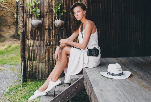 Mujer hermosa joven en vacaciones tropicales en asia, estilo veraniego, vestido blanco boho, zapatillas, cámara de fotos digital, viajero, sombrero de paja, sonriente, boho