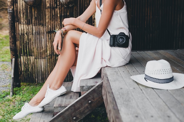 Mujer hermosa joven en vacaciones tropicales en asia, estilo de verano, vestido blanco boho, zapatillas de deporte, cámara de fotos digital, viajero, sombrero de paja, piernas cerca de detalles