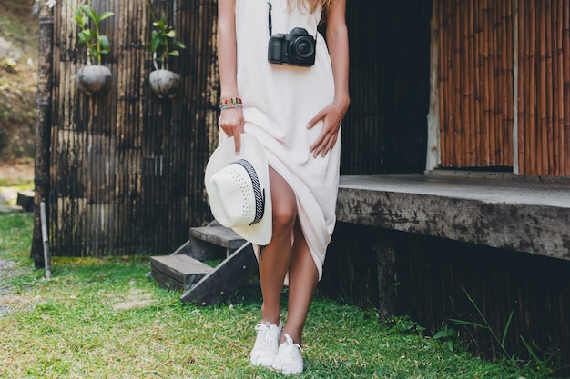 Mujer hermosa joven en vacaciones tropicales en asia, estilo de verano, vestido blanco boho, zapatillas de deporte, cámara de fotos digital, viajero, sombrero de paja, piernas cerca de detalles