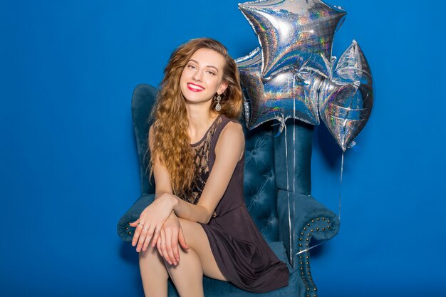 Mujer hermosa joven en vestido gris sentado en un sillón azul con globos plateados