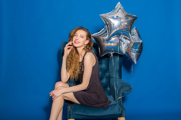 Mujer hermosa joven en vestido gris sentado en un sillón azul con globos plateados