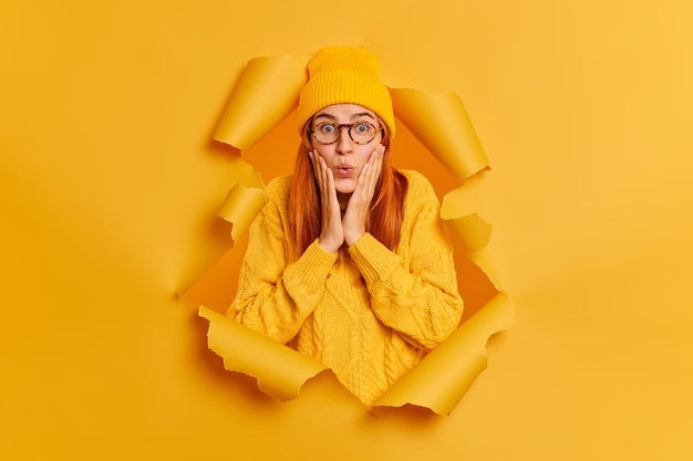 Mujer hermosa pelirroja sorprendida mantiene las manos en las mejillas ve algo increíble mira fijamente viste suéter amarillo y el sombrero se rompe a través del agujero de papel. Mujer impresionada asombrada interior