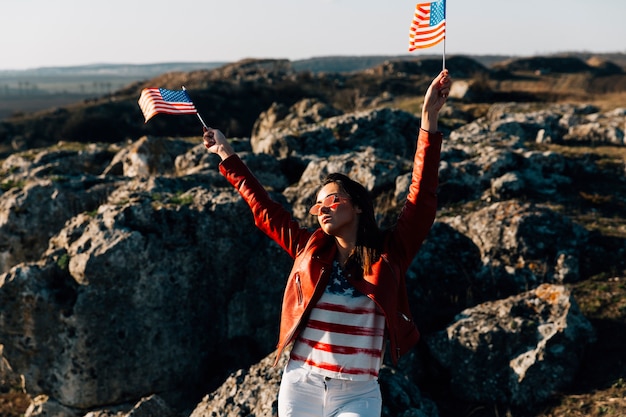 Mujer hermosa que agita banderas americanas en fondo rocoso