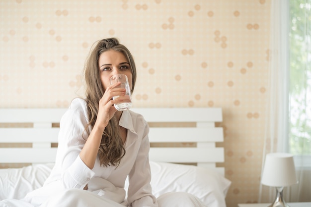 Mujer hermosa que bebe el agua dulce en la cama en la mañana