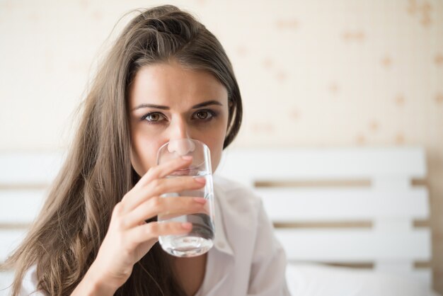 Mujer hermosa que bebe el agua dulce en la cama en la mañana