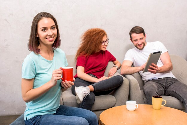 Mujer hermosa que mira la cámara que sostiene la taza de café que se sienta con los amigos que miran la tableta digital