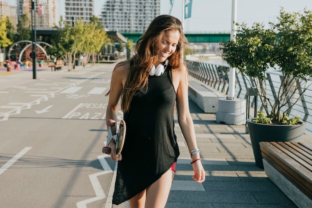 Foto gratuita mujer hermosa que sostiene el monopatín que camina en la calle en ciudad