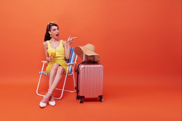 Una mujer hermosa se relaja sentada en una silla de playa con una maleta apuntando con su dedo en el estudio de verano