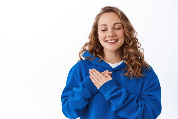 Mujer hermosa soñadora recuerda un momento encantador y sincero, tomados de la mano en el pecho y sonríe con los ojos cerrados, soñando despierto, pensando en algo, pared blanca