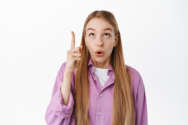 Mujer hermosa sorprendida con el pelo largo y rubio mirando un anuncio en la parte superior, jadeando y diciendo wow, apuntando con el dedo hacia arriba en la promoción, de pie sobre una pared blanca