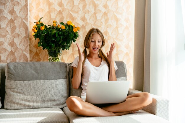 La mujer hermosa vio la sorpresa alegre en la computadora portátil que se sentaba en el sofá en casa