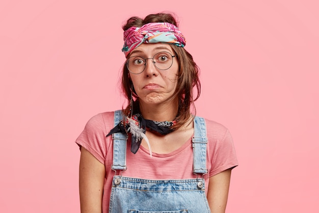 Una mujer hippie bastante dudosa duda si ir de fiesta con amigos, tiene una expresión despistada, frunce los labios, usa gafas redondas