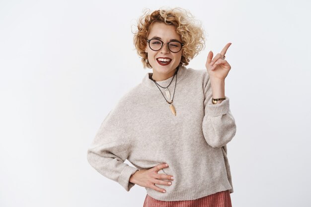 Mujer hipster bastante rubia linda descarada y coqueta con sonrisa carismática guiñando un ojo con alegría y apuntando a la esquina superior derecha