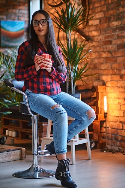 Una mujer hipster morena vestida con una camisa de lana y jeans bebe café en una habitación con interior de loft.