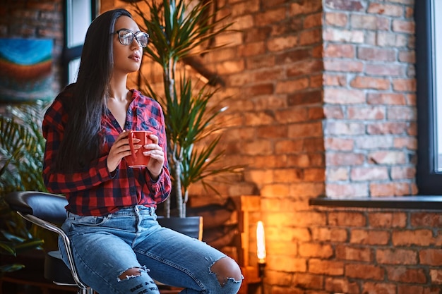 Una mujer hipster morena vestida con una camisa de lana y jeans bebe café en una habitación con interior de loft.