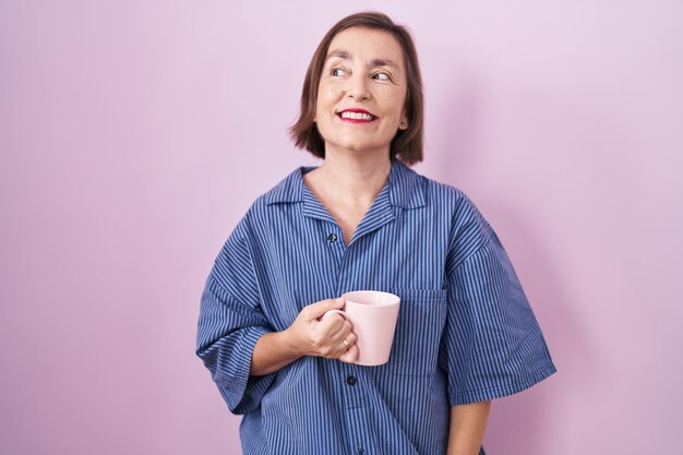 Mujer hispana de mediana edad bebiendo una taza de café sonriendo mirando hacia un lado y mirando hacia otro lado pensando.