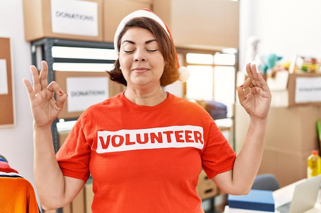 Foto gratuita mujer hispana de mediana edad con camiseta voluntaria y sombrero de navidad relajarse y sonreír con los ojos cerrados haciendo gesto de meditación con los dedos concepto de yoga