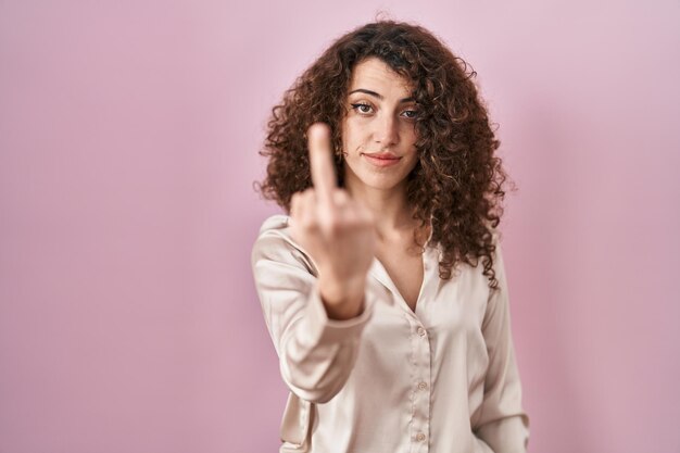 Mujer hispana con el pelo rizado de pie sobre fondo rosa mostrando el dedo medio, expresión descortés y grosera de mierda