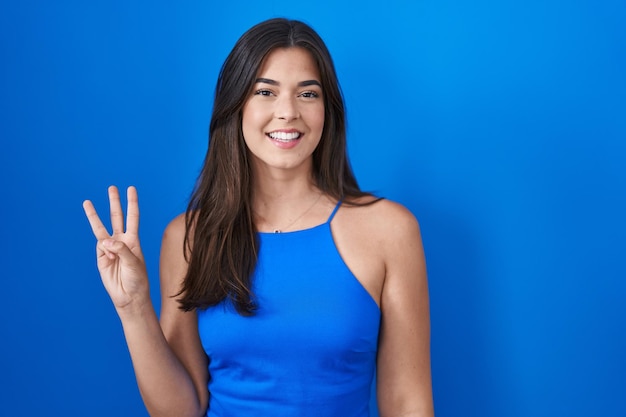 Mujer hispana de pie sobre fondo azul que aparece y señala con los dedos número tres mientras sonríe confiada y feliz.