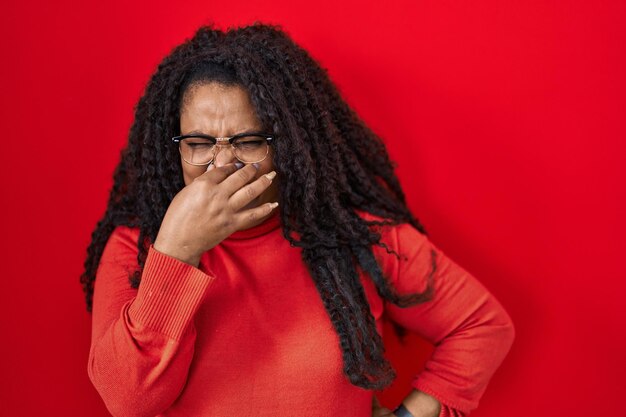 Mujer hispana de talla grande parada sobre fondo rojo oliendo algo apestoso y repugnante olor intolerable aguantando la respiración con los dedos en la nariz mal olor