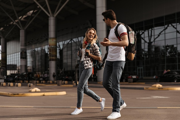 Mujer y hombre alegres con camisetas blancas y jeans caminan y hablan cerca del aeropuerto Retrato completo de viajeros con mochilas
