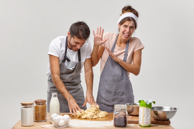 Foto gratuita la mujer y el hombre divertidos hacen masa para pastel, tienen caras alegres, se visten con delantales, obtienen experiencia culinaria, tienen algunos problemas, agregan ingredientes que no están de acuerdo con la receta. tiempo de cocción, concepto de horneado