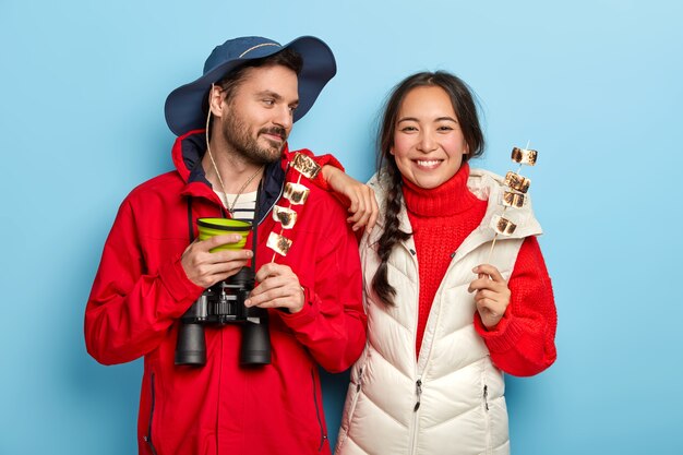 La mujer y el hombre felices pasan el tiempo libre en la naturaleza, disfrutan de la actividad de campamento, sostienen malvaviscos asados, toman café, comen y beben juntos, vestidos con ropa informal