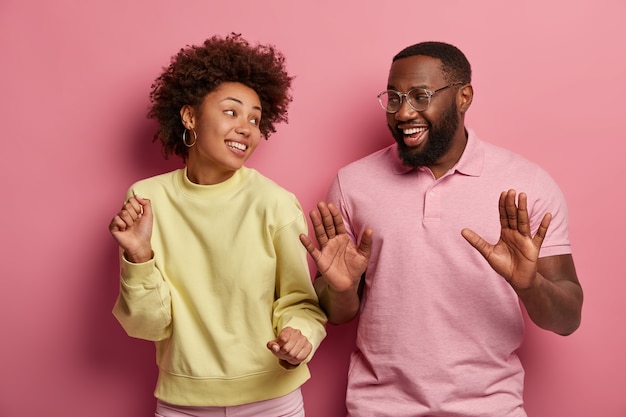 Mujer y hombre milenarios llenos de alegría, sonríe ampliamente, baila con las manos en alto, disfruta del ritmo de la música, usa ropa informal