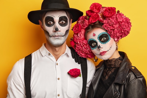 La mujer y el hombre muertos llevan maquillaje de calaveras, pintados para Halloween, miran a la cámara sorprendentemente, vestidos con traje blanco y negro para Todos los Santos, aislado sobre fondo amarillo.