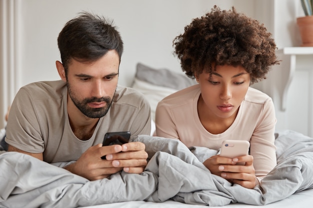 La mujer y el hombre multiétnicos adictos buscan información en la red, usan teléfonos celulares modernos, miran videos o leen blogs, descansan en la cama, usan internet inalámbrico. Tecnologías modernas y adicción