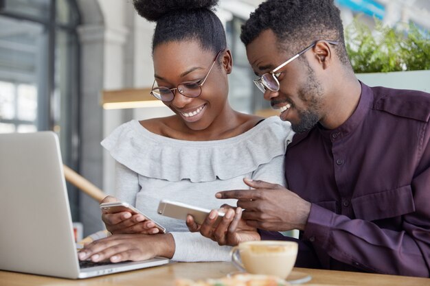 La mujer y el hombre negros tienen una reunión informal, me alegra ver fotos en un teléfono inteligente, usar gafas, trabajar juntos en un proyecto común a través de una computadora portátil