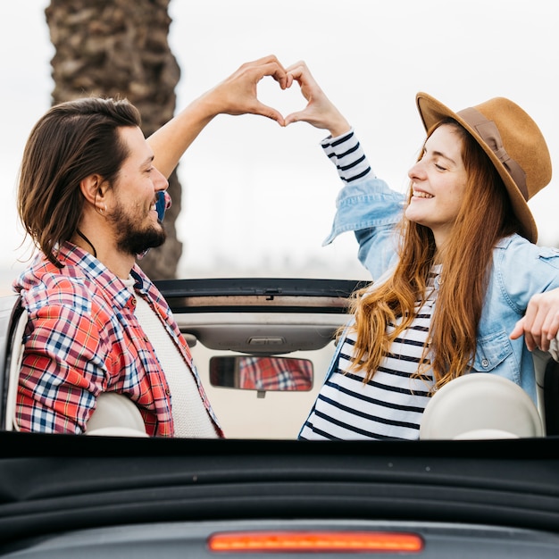 Mujer y hombre sonrientes jovenes que muestran el símbolo del corazón y que se inclinan hacia fuera del coche