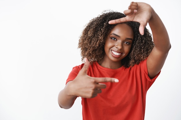 mujer imaginando cómo aplicar oportunidades a la vida, imaginando la imagen como haciendo un marco con los dedos y mirando a través de ella sonriendo sintiéndose creativa y optimista posando encantada sobre una pared blanca