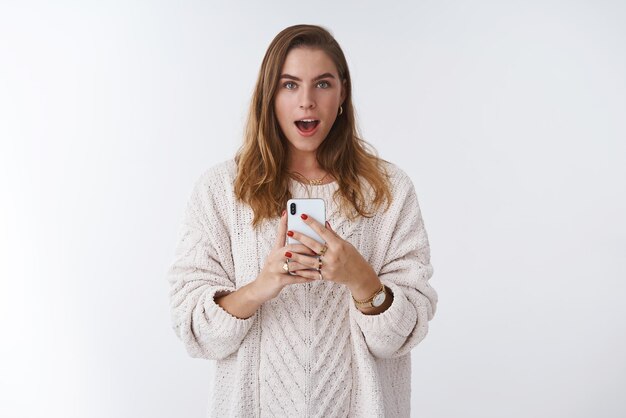 Mujer imitando mirando descarado celebración smartphone tomando selfie espejo boca abierta divertido de pie blanco vistiendo suelto suéter de moda accessorize