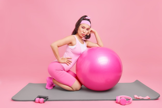 mujer se inclina hacia fitball inflado tiene entrenamiento fintess en casa gimnasio ha peinado cola de caballo usa ropa deportiva lleva un estilo de vida saludable aislado en rosa