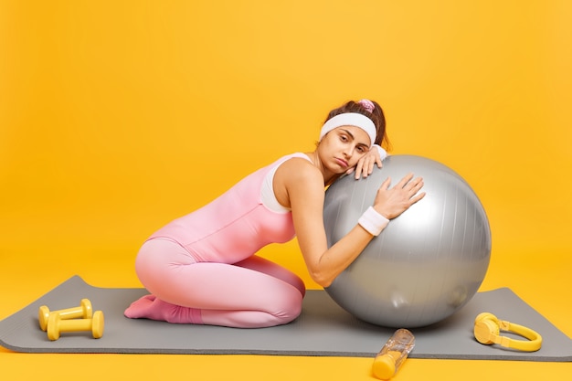 La mujer se inclina hacia la pelota de fitness siente fatiga después de que el entrenamiento aeróbico usa una diadema con muñequera y poses de ropa deportiva en karemat aislado en amarillo