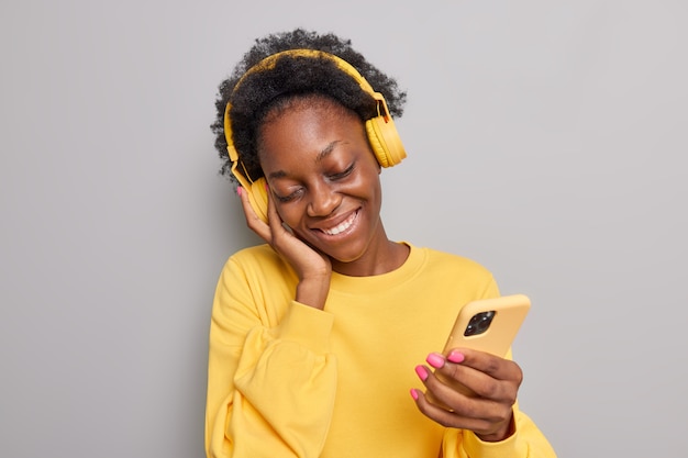 Mujer se inclina sonríe complacido escucha música en auriculares inalámbricos tiene smartphone vestido con puente amarillo aislado en gris