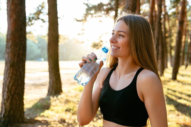 Mujer increíble alegre bebiendo agua de botella después del entrenamiento en el parque, al aire libre