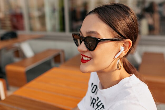 mujer increíble con una amplia sonrisa y labios rojos con gafas de sol oscuras y camiseta blanca sentada en la terraza de verano