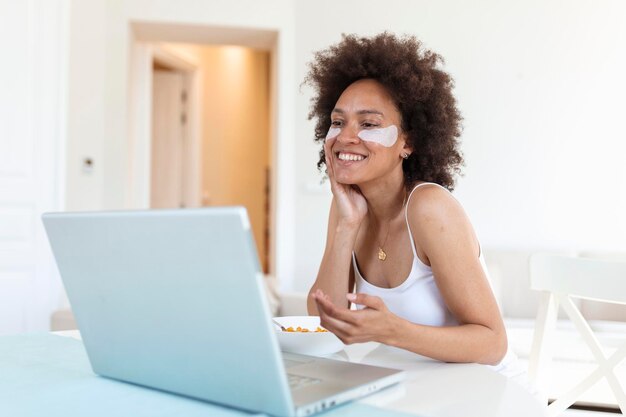 Mujer increíble joven sentada en el interior en la mesa con una computadora portátil sosteniendo copos de maíz Mirando la computadora portátil y hablando con sus amigos a través de una videollamada