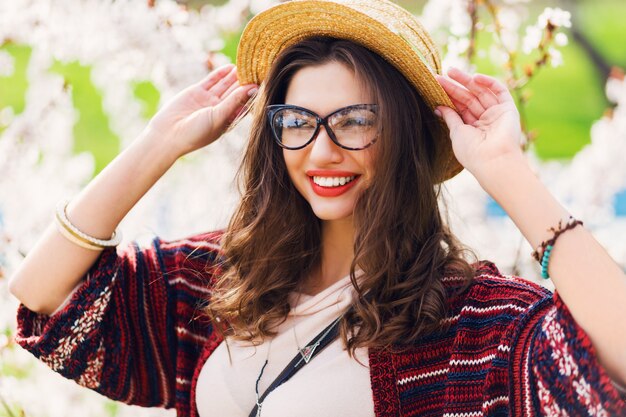 Mujer increíble con maquillaje brillante, ojos azules, gafas, sombrero de paja posando en el parque soleado de primavera cerca del árbol de flores