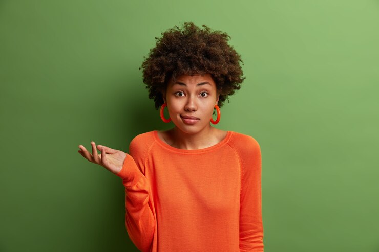 Foto gratuito la mujer indecisa dudosa levanta la palma de la mano con vacilación, se enfrenta a una pregunta difícil o dos opciones, usa un suéter naranja y aretes, aislados en la pared verde. personas, percepción y actitud