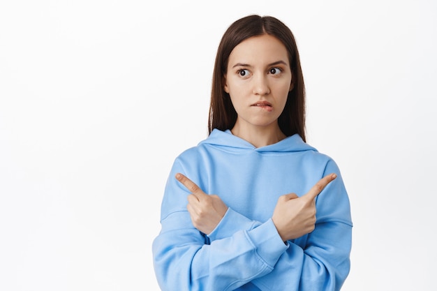 Mujer indecisa tratando de elegir entre dos variantes, apuntando de lado a dos pancartas de venta, mordiéndose el labio pensativo, pensando, haciendo elección, de pie sobre una pared blanca