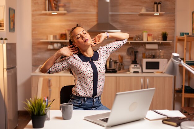 Mujer independiente estirando los brazos debido al agotamiento mientras trabaja horas extras desde casa. Empleado que usa tecnología moderna a medianoche haciendo horas extras para trabajo, negocios, ocupado, carrera, red, estilo de vida