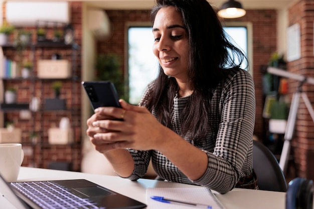 Mujer independiente que trabaja en una aplicación para teléfonos inteligentes y usa una computadora portátil para un trabajo remoto en el escritorio de su casa. Navegación por Internet y redes sociales en teléfonos móviles a mensajes de texto, descanso de investigación en línea.