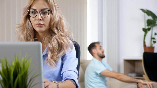 Mujer independiente que trabaja en la computadora portátil de la casa mientras el esposo ve la televisión en el fondo